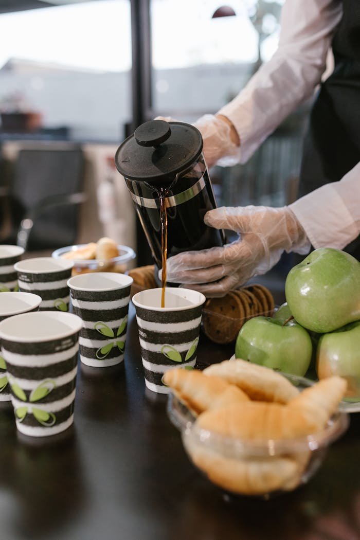 hero-img-02 Barista pours coffee from French press into cups at a breakfast event with croissants and apples.